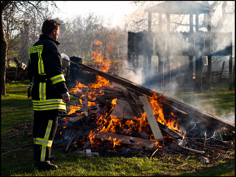 Foto: brennende Holzscheite, Feuerwehr überwacht das Ganze