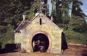 Gemälde: Hufschmiede in Irland am Waldrand, Backsteinbau mit einem als Hufeisen geformten Tor, innen Mann mit Pferd und Hufschmied