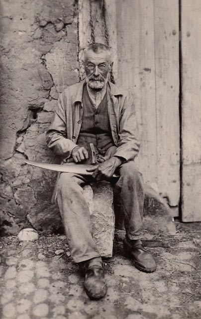 altes sw-Foto: alter Mann mit Nickelbrille sitzt auf einem Stein und beschlägt Blech mit einem Hammer