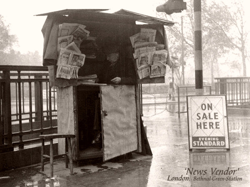 sw-Foto: älterer Mann sitzt unter einer Holzbuden ähnlichen Überdachung, neben ihm Zeitungsaushänge zum Verkauf, aber kein Käufer in Sicht - es regnet ...