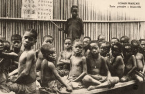 s/w Foto: stehender Junge, der mit Zeigestock auf Pappschild mit Alphabet und Ziffern weist, zwischen mehreren sitzendenSchulanfängern