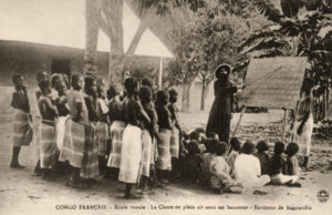 s/w Foto: im Freien unter Banananstaude Lehrer an einer Stehtafel, links davor sitzende und stehende Schüler