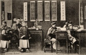 s/w Foto: chinesischer Lehrer an einem Sitzpult mit mehreren Schülern in einem Klassenzimmer