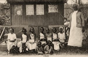 s/w Foto: im Freien stehende Lehrerin mit Zeigestock auf als Tafel dienende Bretterwand weisend, davor neun zum Foto aufgereiht sitzende Schülerinnen und Schüler