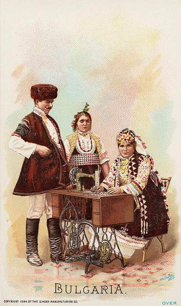 alte Postkarte: eine Frau sitzt an Nähmaschine, Paar steht daneben, alle in Tracht