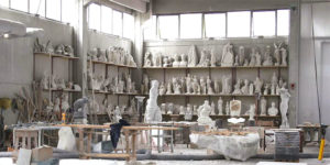 Farbfoto: Blick in eine Bildhauerwerkstatt mit Werkzeugen, Steinblöcken und vielen Skulpturen und Büsten in Regalen an den Wänden