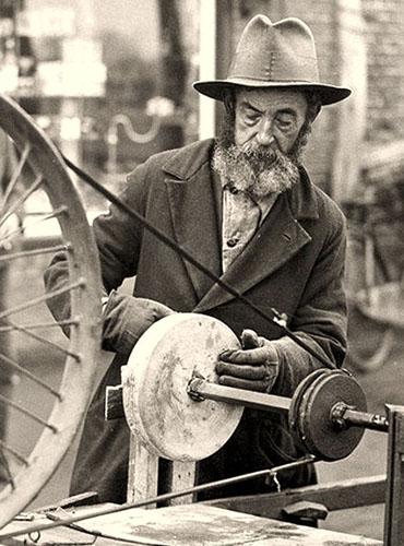 s/w Foto: alter Scherenschleifer mit Hut und Bart am Straßenrand bei der Arbeit am Schleifstein