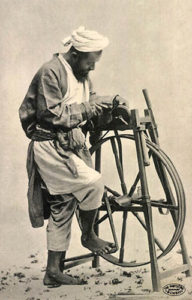 Scherenschleifer, Messerschleifer alte s/w Fotopostkarte: Inder mit Turban bedient im Sand stehendes Schleifgerät mit einer Fußkurbel, während er ein Messer schärft