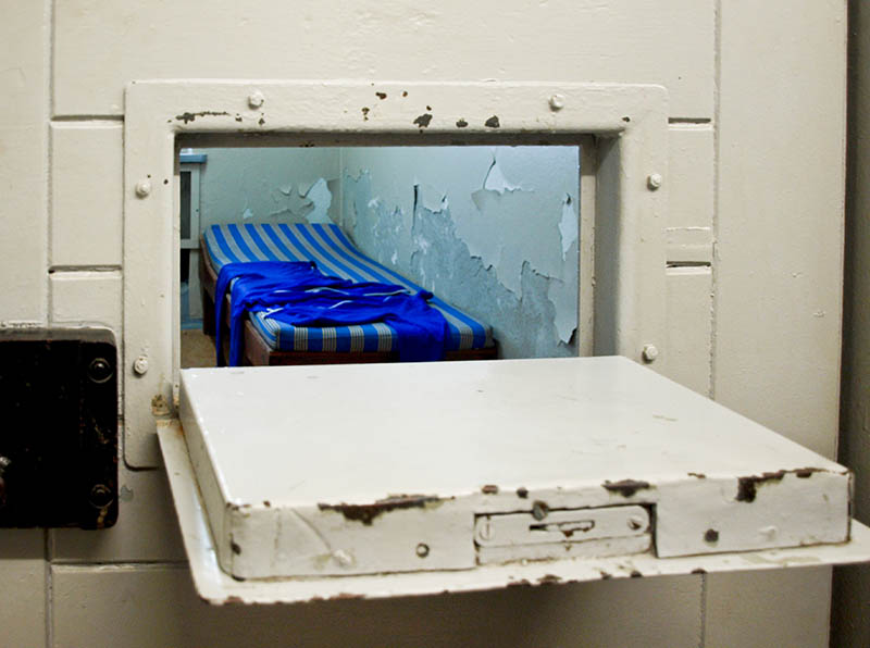 Farbfoto: Einblick durch geöffnete Gefängnistürklappe in die Zelle. Dort steht ein einfaches Bett mit Matratze.