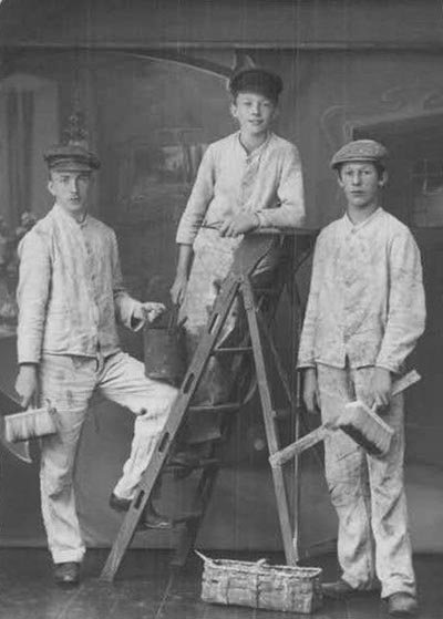 sw-Foto: drei Jugendliche in Malerkleidung mit Malerpinsel; einer von ihnen steht auf einer Leiter