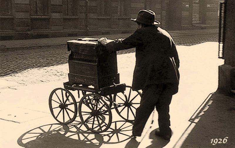 altes s/w Foto: alter Mann mit Leierkasten auf vier Rädern auf Kopfsteinpflasterstraße im Gegenlicht