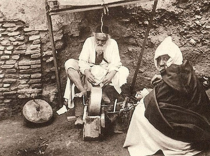 altes s/w-Foto: jenischer Messerschleifer sitzt am Schleifstein am Boden, davor sitzt wohl ein Kunde