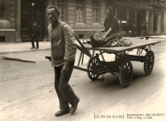 sw Foto: Mann zieht mittels Deichsel und Schultergurt Pritschenwagen, nebst darauf sitzender Frau