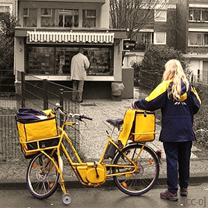 Farbfoto: Zustellerin mit Postrad am Straßenrand