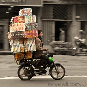Farbfoto: Lieferant mit großem Paketstapel auf Moped unterwegs