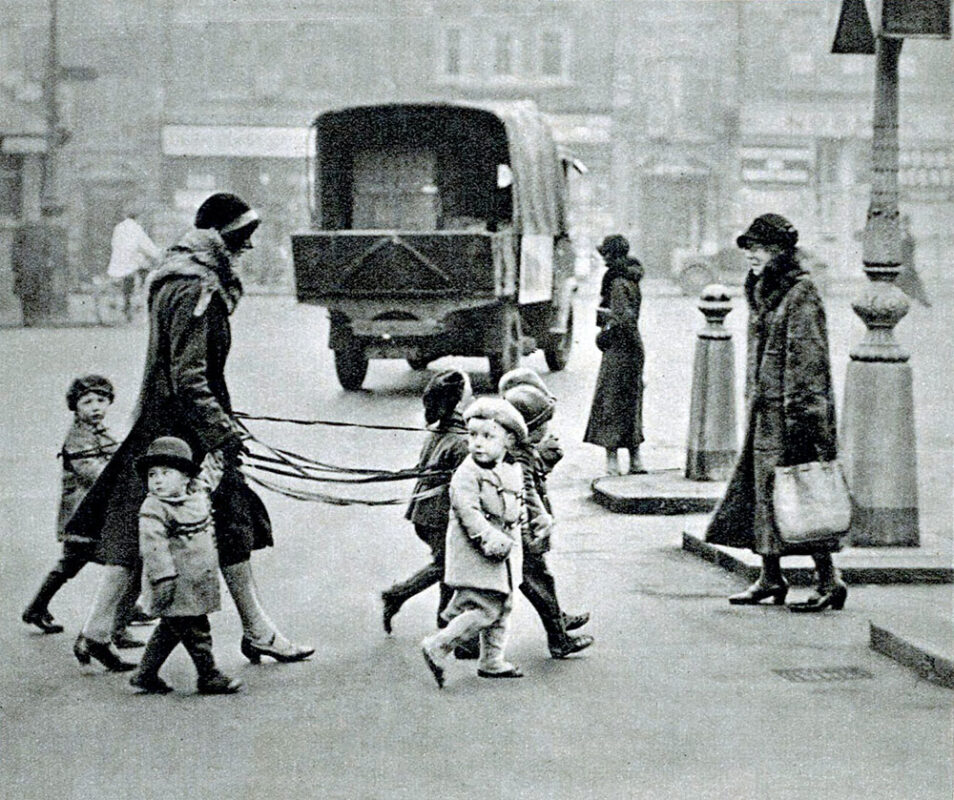 Frau führt mehrere Kleinkindern mit Bändern über die Straße